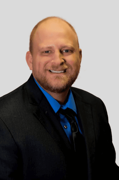 Matt-MMcLean-Headshot A man with a reddish beard and short hair is wearing a black suit jacket over a blue shirt and black tie, smiling at the camera against a plain light background.